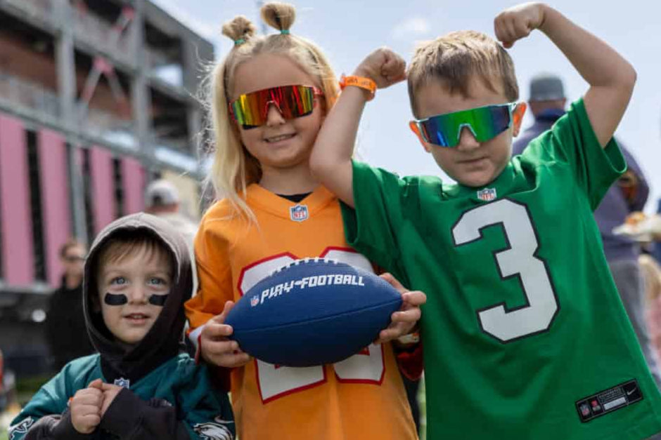 Three children in sports jerseys and sunglasses holding a football on a sports field.