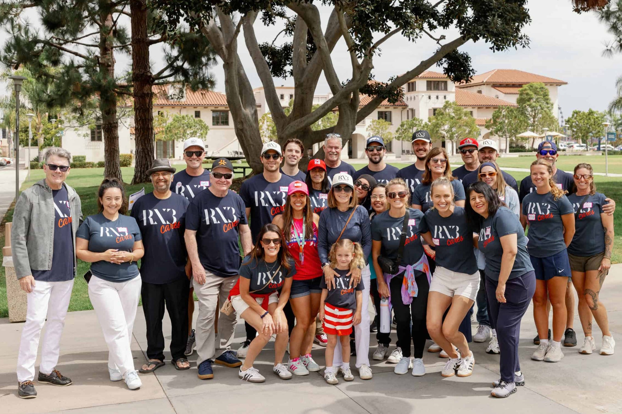 Group of people wearing navy blue shirts with 'RX3' logo outdoors on a sunny day.